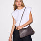 A woman wearing a white sleeveless top and dark jeans models a brown leather shoulder bag with gold-tone studs and hardware against a plain white studio background.