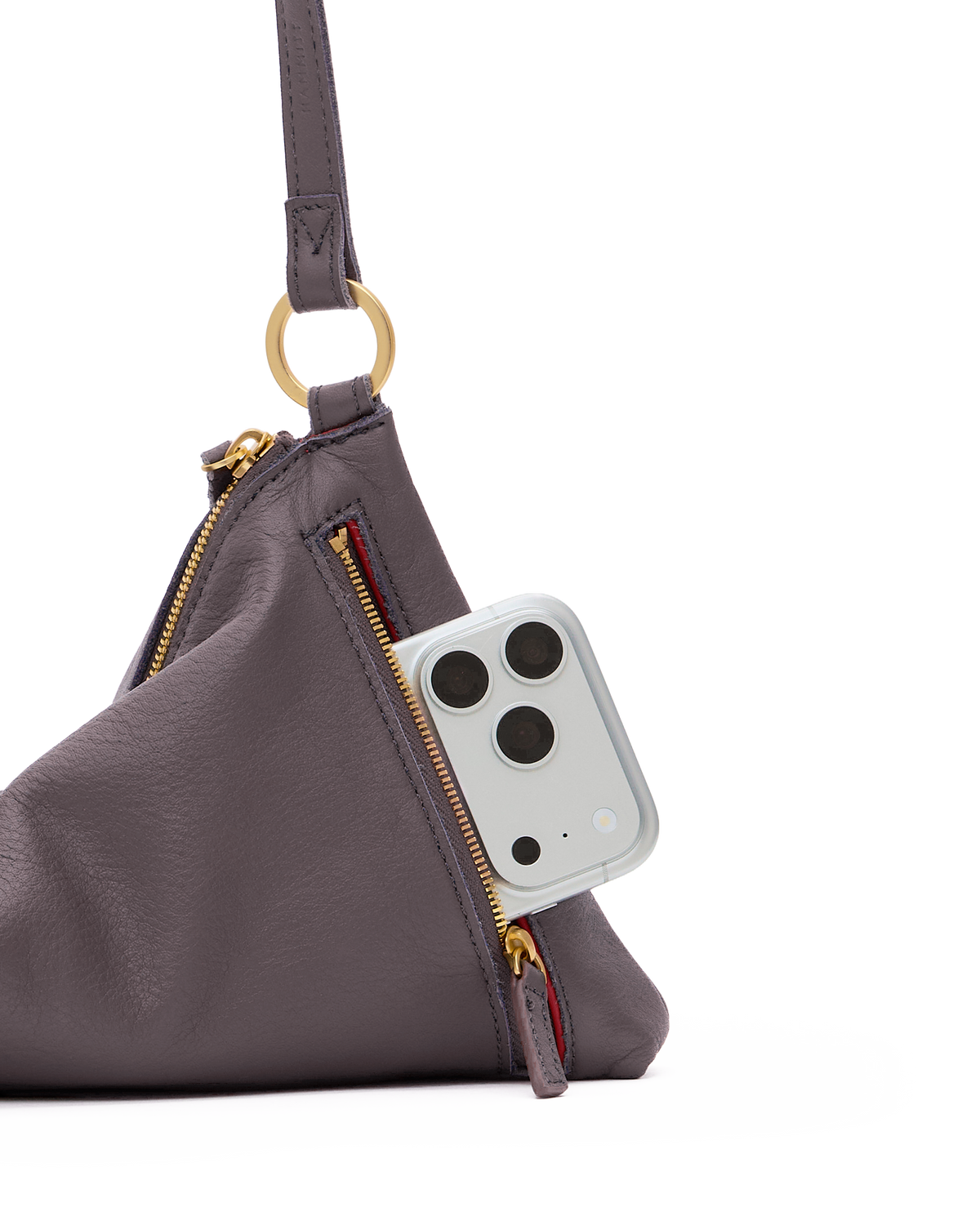A close-up studio shot of a dark gray pebbled leather shoulder bag with gold-tone hardware, including a ring handle and zipper pulls. A light gray smartphone is tucked into the side zipper pocket of the bag, set against a clean white background.
