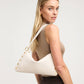 A blonde model wearing a beige tank top and dark pinstriped trousers carries a large off-white shoulder bag with gold-tone hardware and a white strap against a light gray studio background.