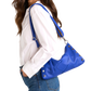 A woman wearing a white button-up shirt and blue jeans carries a bright blue leather shoulder bag with silver-tone studs and hardware.