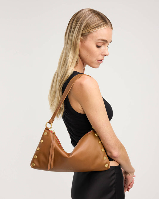 A blonde woman in a black sleeveless top and black skirt carries a tan leather shoulder bag with gold-tone studs and a matching strap against a light gray studio background.