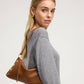 A blonde woman wearing a gray knit sweater and gold hoop earrings carries a brown leather shoulder bag with gold-tone studs and a matching strap against a light gray studio background.