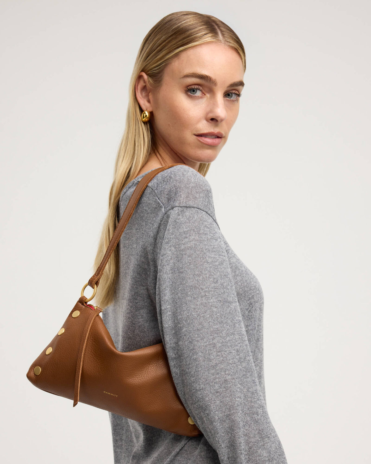 A blonde woman wearing a gray knit sweater and gold hoop earrings carries a brown leather shoulder bag with gold-tone studs and a matching strap against a light gray studio background.