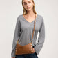 A woman wearing a gray long-sleeve V-neck sweater and blue jeans, carrying a brown leather shoulder bag with gold-tone hardware and a matching strap over her shoulder, against a light gray studio background.