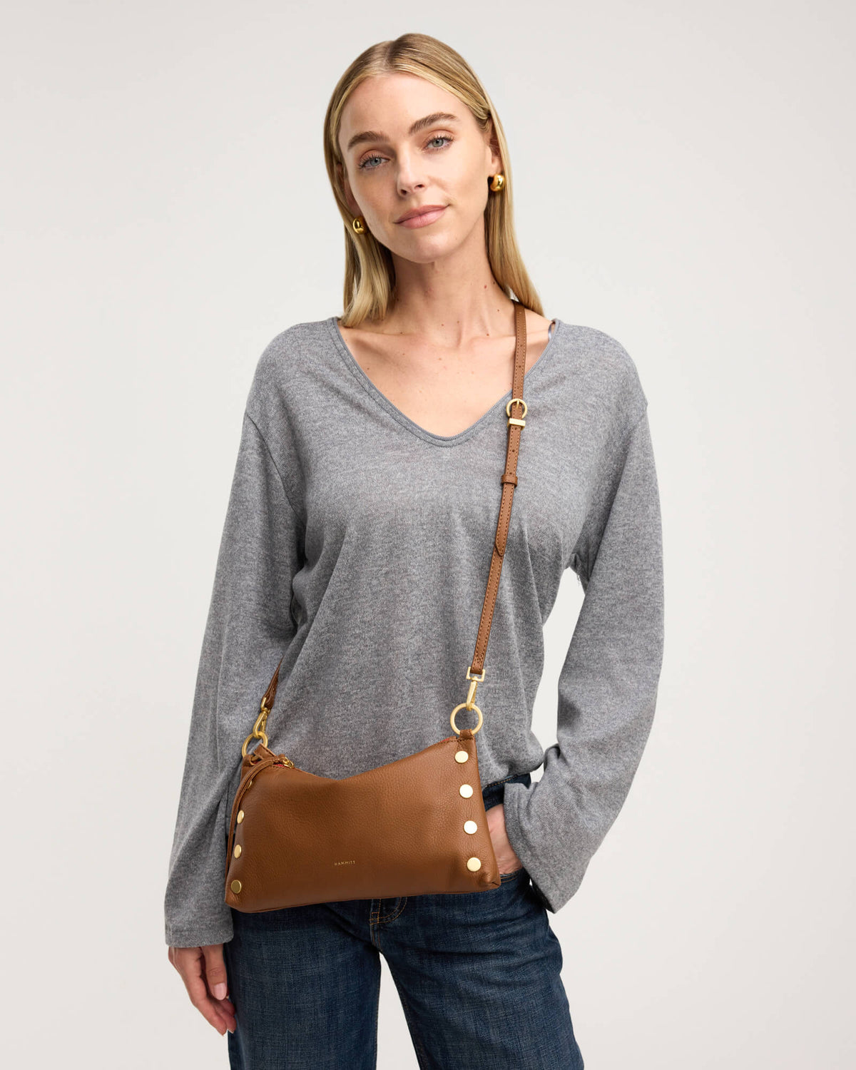 A woman wearing a gray long-sleeve V-neck sweater and blue jeans, carrying a brown leather shoulder bag with gold-tone hardware and a matching strap over her shoulder, against a light gray studio background.