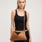 A woman wearing a black ribbed tank top and black skirt models a brown leather shoulder bag with gold-tone hardware and a matching brown belt. The bag features a slouchy triangular shape with gold studs along the edges and a thin adjustable strap.