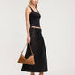 A woman modeling a black sleeveless crop top and a black satin midi skirt, holding a brown leather shoulder bag with gold-tone hardware, against a light gray studio background.