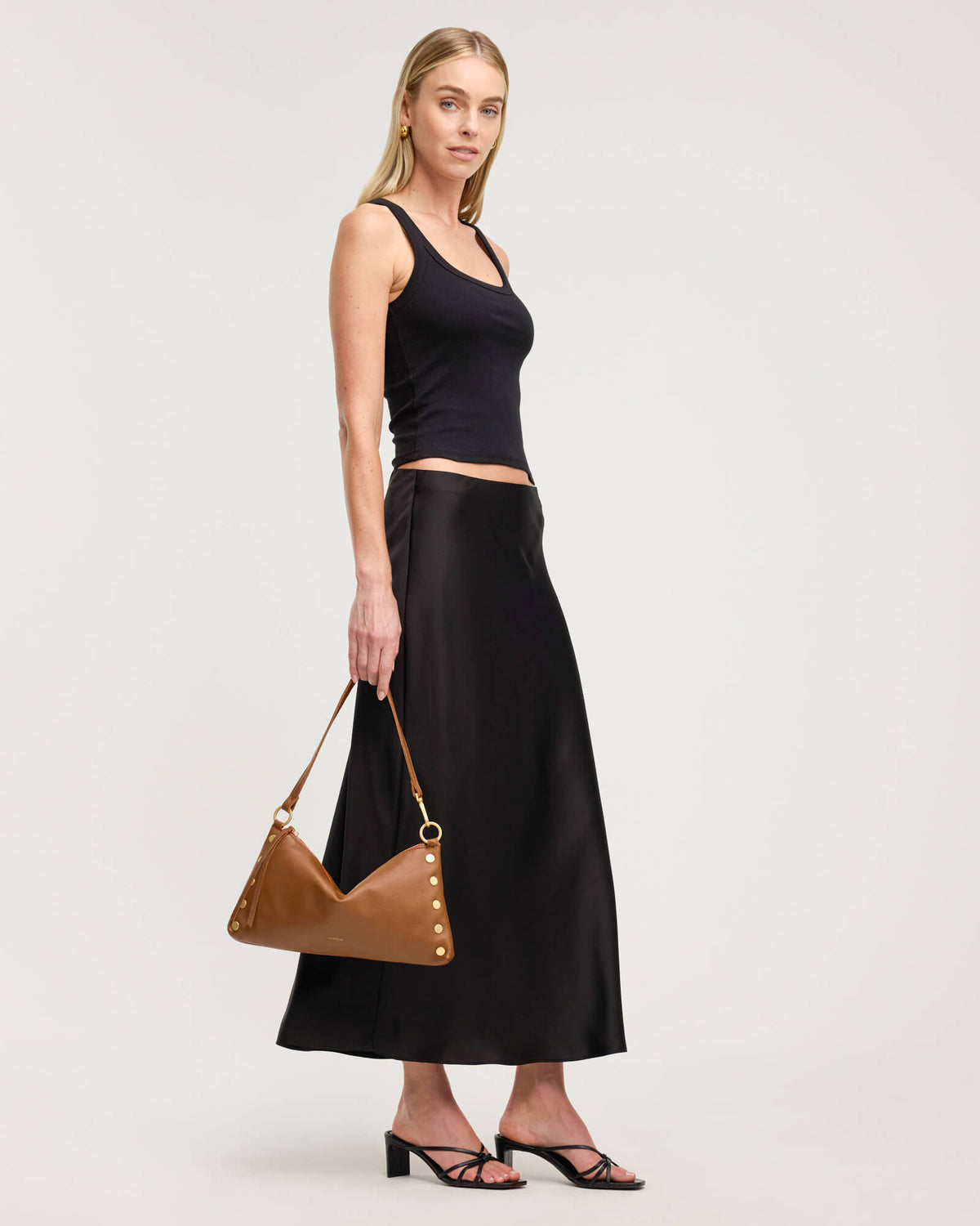 A woman modeling a black sleeveless crop top and a black satin midi skirt, holding a brown leather shoulder bag with gold-tone hardware, against a light gray studio background.