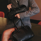 A model in a gray pinstripe blazer holds two black leather handbags, one larger satchel-style bag and one structured briefcase-style bag.