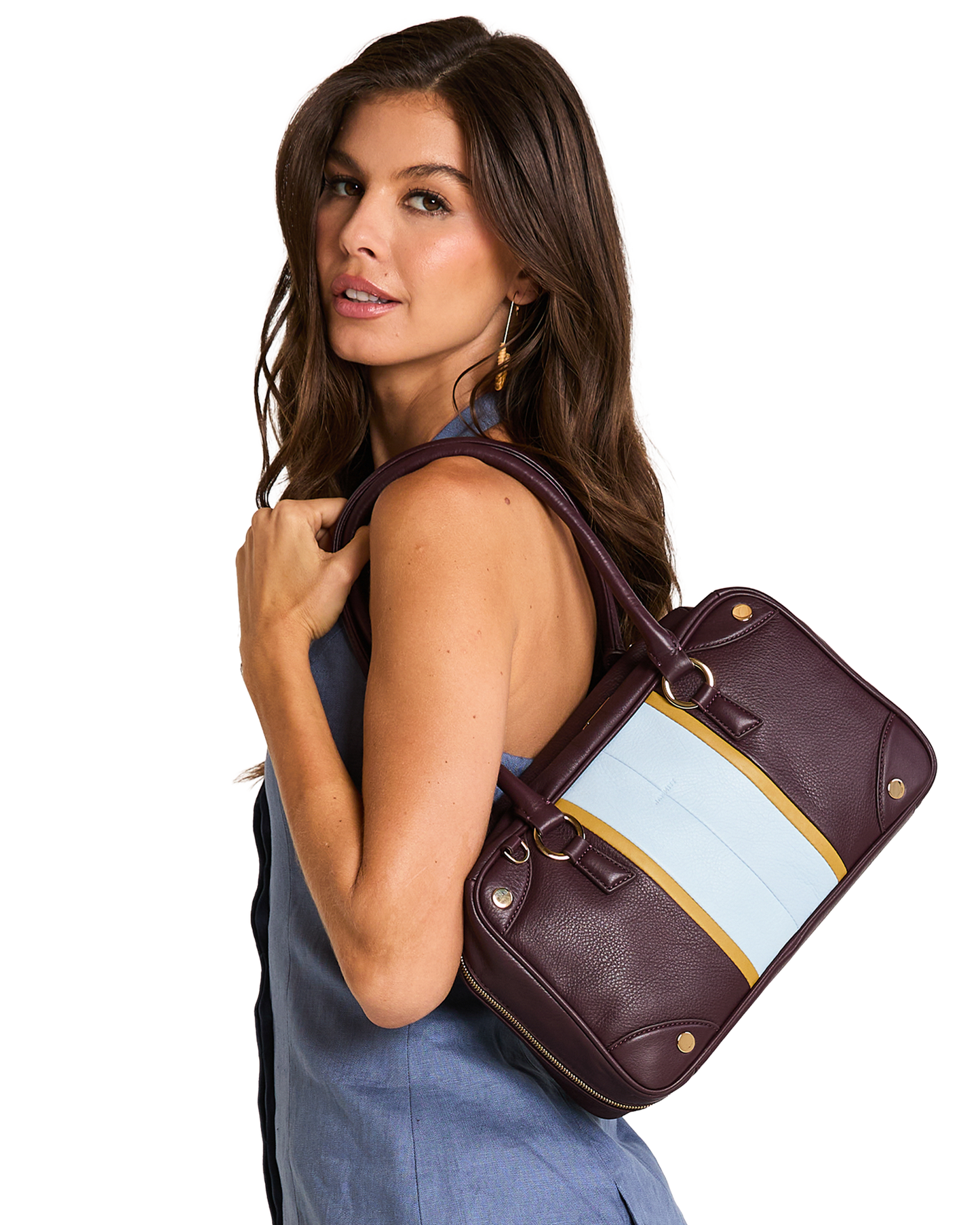 A model wearing a blue sleeveless top carries a structured leather handbag over her shoulder. The bag is deep burgundy with a wide light blue panel and a thin gold stripe detail.