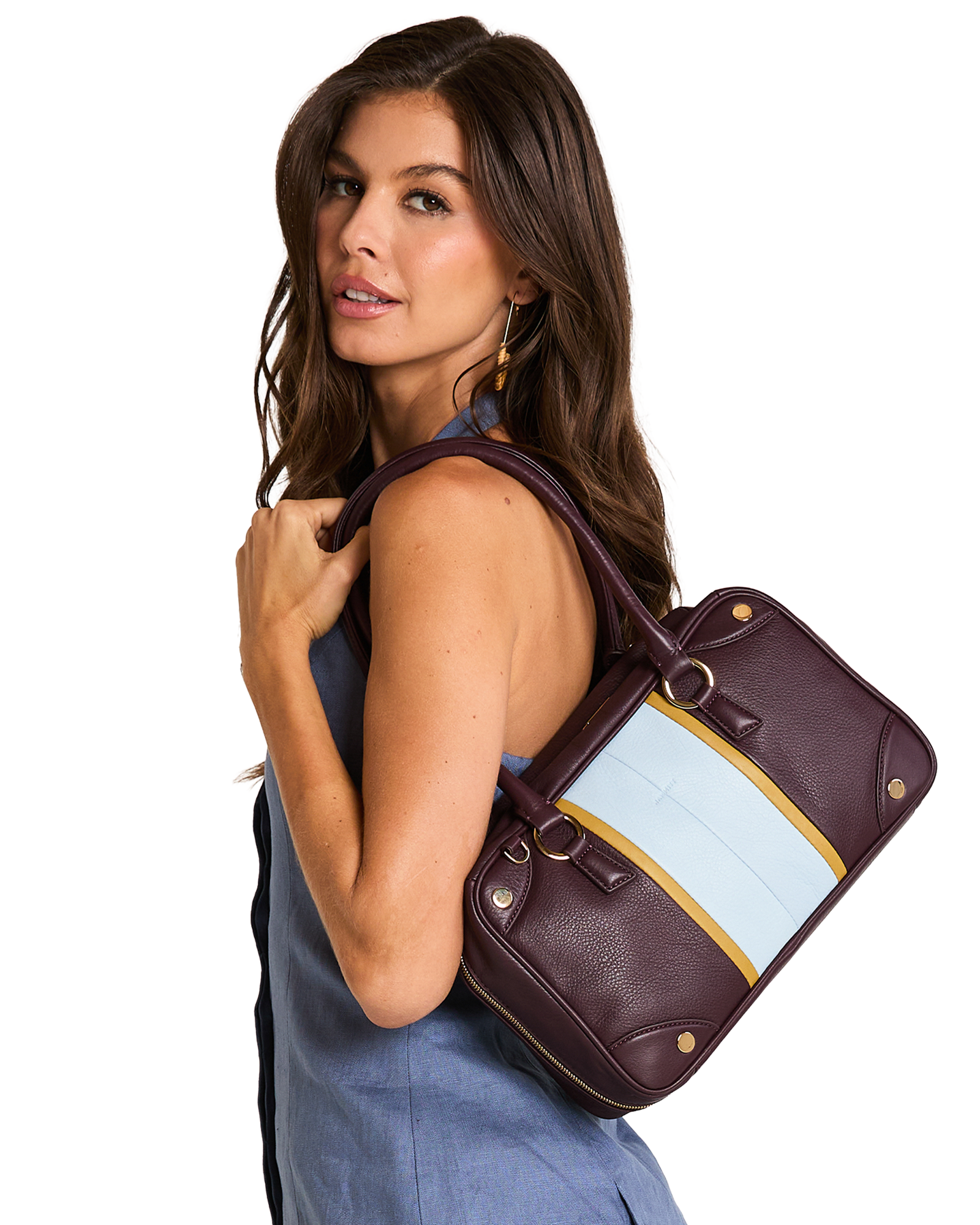 A model wearing a blue sleeveless top carries a structured leather handbag over her shoulder. The bag is deep burgundy with a wide light blue panel and a thin gold stripe detail.