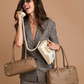 A woman in a gray plaid blazer and shorts holds two taupe leather handbags, one on each shoulder.