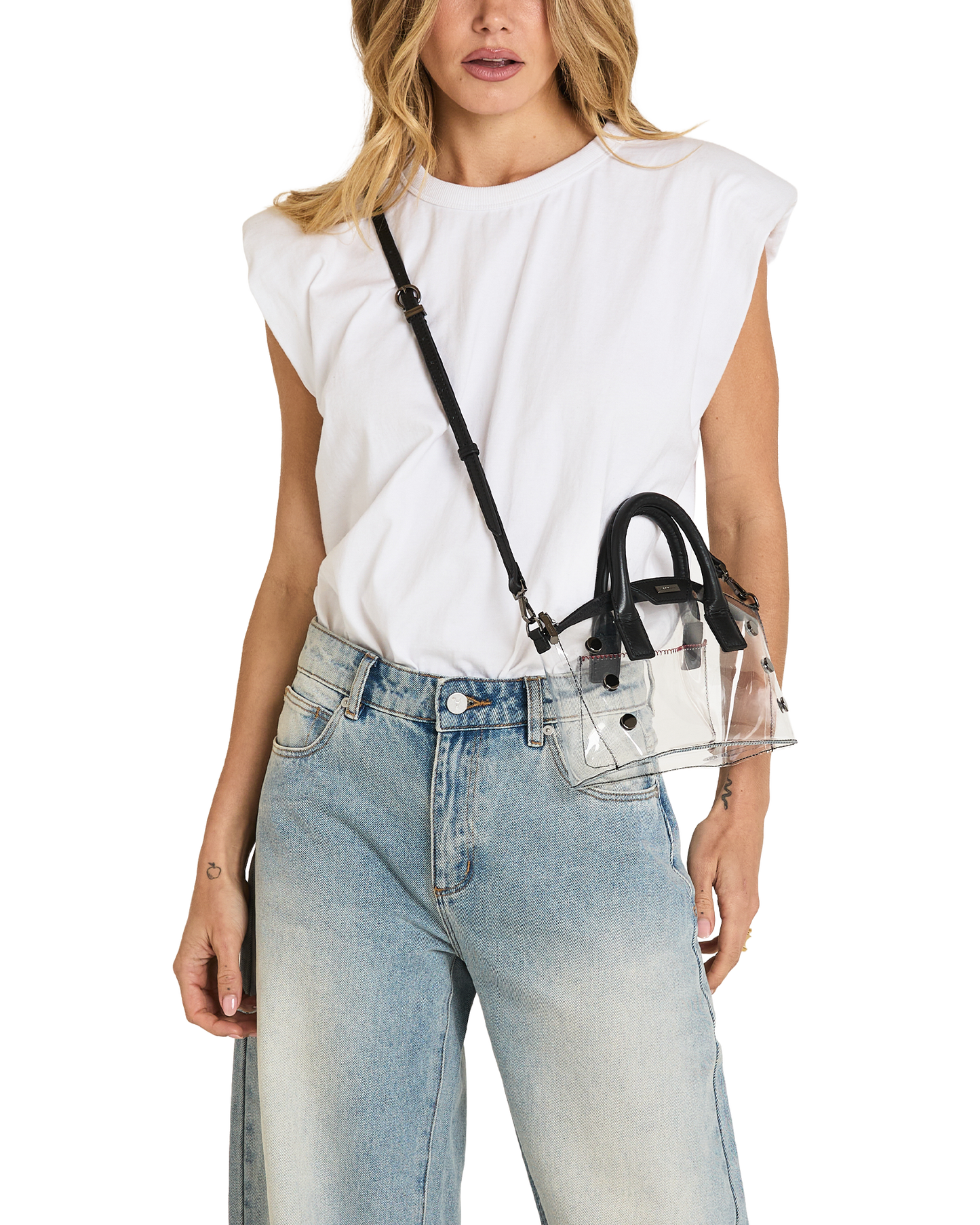A woman wearing a white sleeveless top and light blue jeans carries a small clear handbag with black handles and a black shoulder strap against a plain white background.