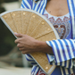 A close-up lifestyle shot of a person holding a woven straw folding fan. The person is wearing a blue and white striped button-up shirt over a white tank top with a red and blue paisley print.