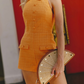 A woman wearing a bright orange textured halter mini dress with button details and flap pockets, holding a woven beige clutch with gold studs against a red and white background.