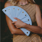 A woman in a beige knit dress holds a light blue faux-leather clutch bag with gold-tone studs and a scalloped edge.
