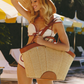 A woman in a white bikini and straw hat carries a large woven straw tote bag with brown leather trim in an outdoor poolside setting.