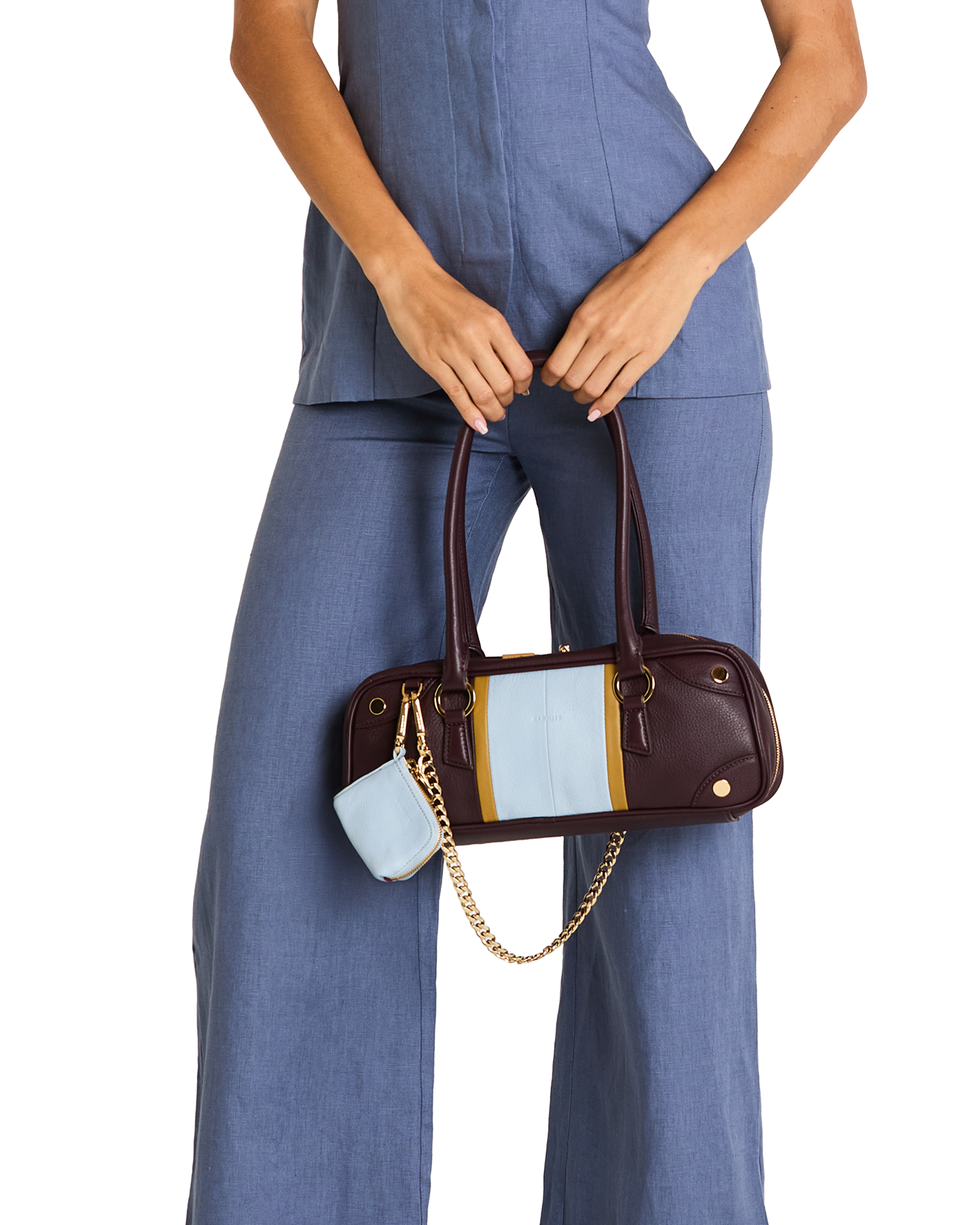 A model wearing a blue jumpsuit holds a dark brown leather handbag with a light blue and yellow color-blocked front panel and a gold chain strap. A small matching light blue pouch hangs from the bag.