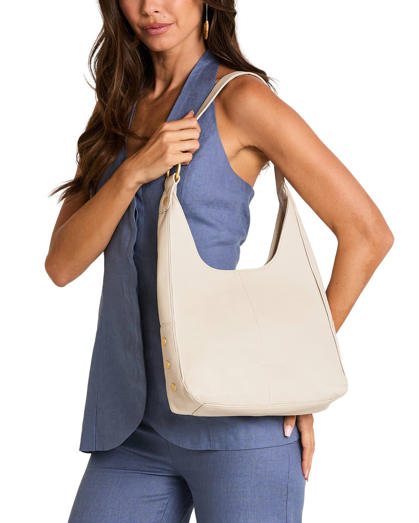 A woman wearing a blue sleeveless top and matching pants carries a large cream-colored leather hobo shoulder bag with gold-tone hardware.