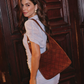 A woman wearing a white puff-sleeve mini dress carries a large brown quilted leather shoulder bag with a quilted pattern and a single strap, standing in front of a dark wood paneled door.