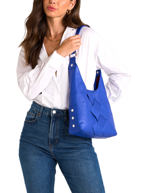 A woman wearing a white button-down shirt and blue jeans carries a bright blue leather shoulder bag with a woven pattern on the front.