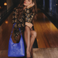 A woman sits on a wooden ledge in a dimly lit indoor setting, wearing a black and gold floral patterned dress and holding a bright blue woven leather shoulder bag.