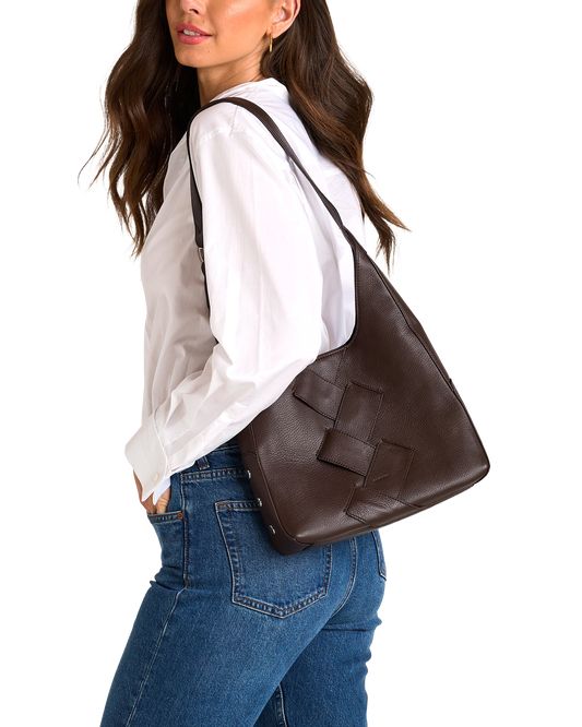 A woman wearing a white blouse and blue jeans carries a large dark brown woven leather shoulder bag against her back.
