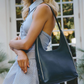 A woman wearing a light blue pinstripe dress carries a large navy blue leather shoulder bag with white piping and silver-tone studs, standing in front of a white-framed window.