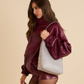 A woman in a matching burgundy faux-leather tracksuit holds a large light gray tote bag with gold-tone buttons and a white shoulder strap, posed against a solid beige studio background.