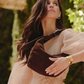A woman outdoors wearing a large brown suede crescent shoulder bag across her torso, paired with a sheer blush dress and gold jewelry.