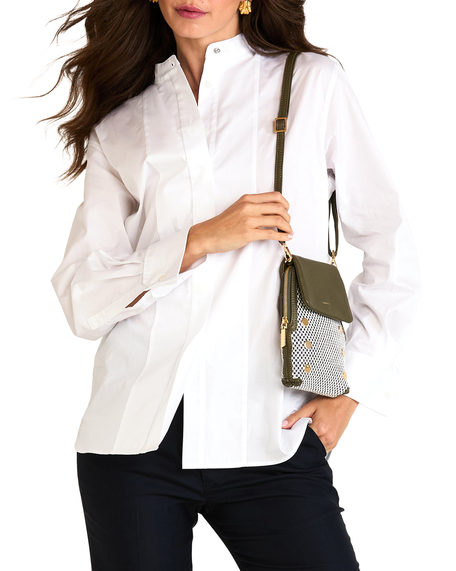 A woman wearing a white button-up blouse and dark pants, holding a small olive green and patterned crossbody bag against a plain white background.
