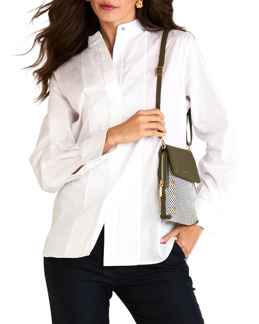 A woman wearing a white button-up blouse and dark pants, holding a small olive green and patterned crossbody bag against a plain white background.
