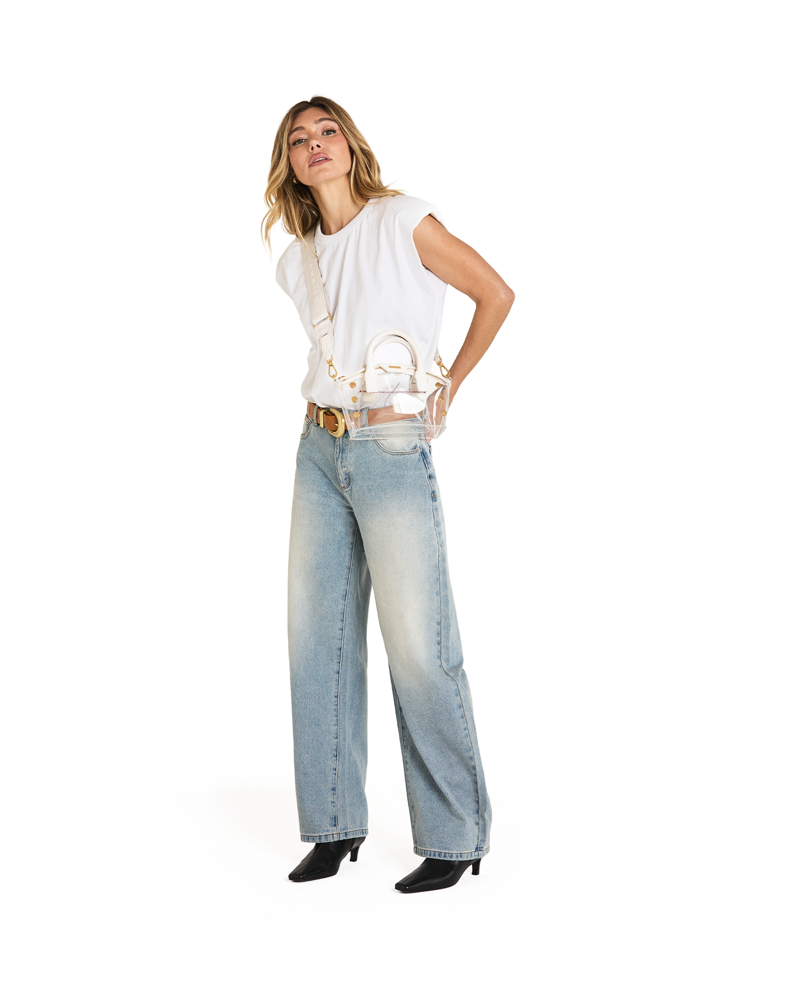 A woman modeling a white sleeveless top, light-wash wide-leg jeans, a gold belt, and black pointed-toe heels, while carrying a small white handbag with a gold chain strap.
