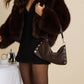 Person wearing a dark fur coat holding a brown leather handbag in a neutral setting.