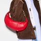 A person wearing a brown suede jacket, light blue button-up shirt, white tee, and blue jeans carries a bright red crescent-shaped shoulder bag with gold-tone hardware against a plain white background.