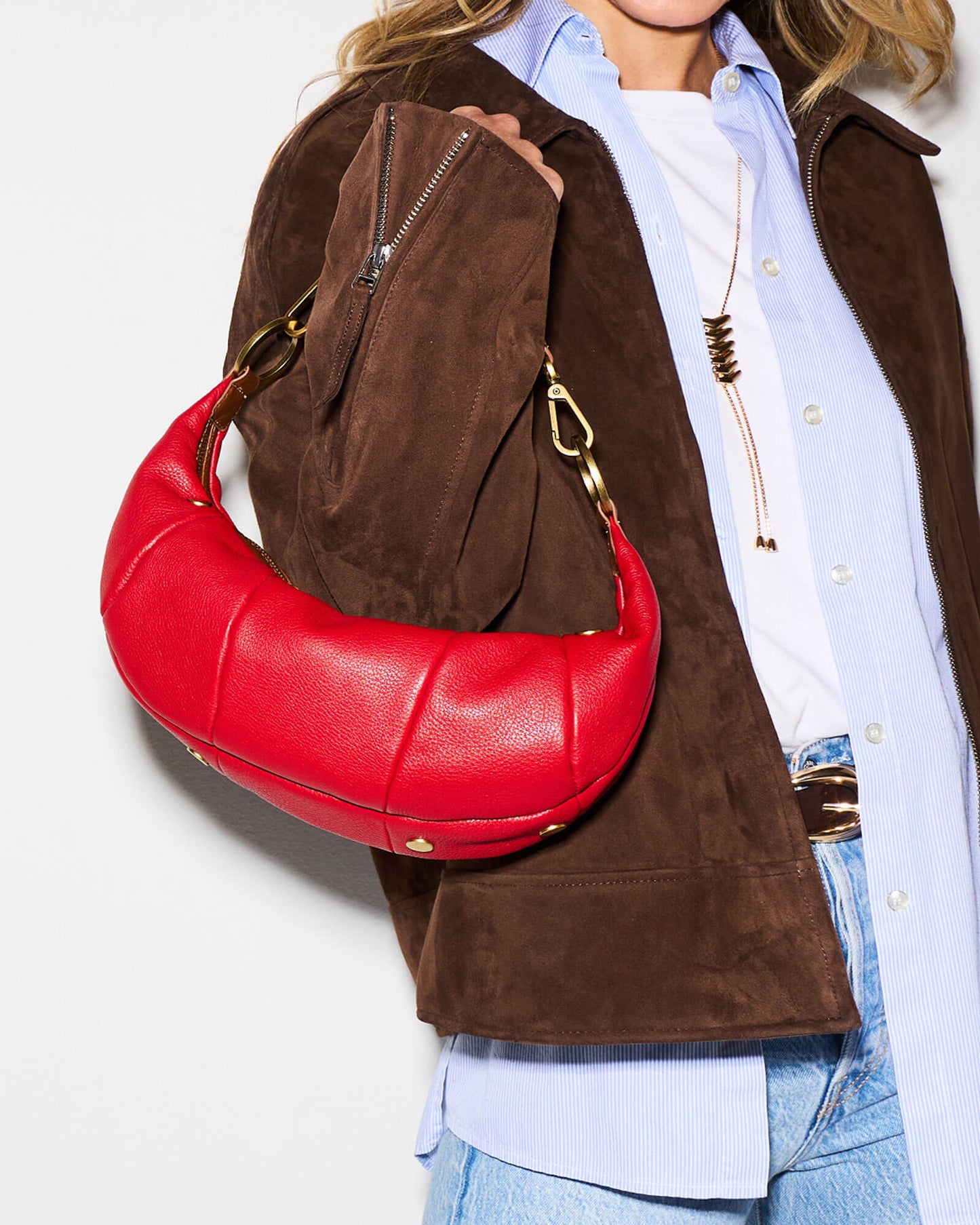 A person wearing a brown suede jacket, light blue button-up shirt, white tee, and blue jeans carries a bright red crescent-shaped shoulder bag with gold-tone hardware against a plain white background.