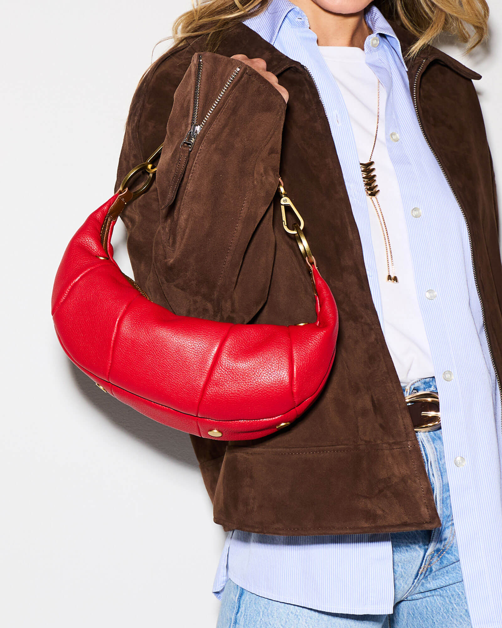 A person wearing a brown suede jacket, light blue button-up shirt, white tee, and blue jeans carries a bright red crescent-shaped shoulder bag with gold-tone hardware against a plain white background.