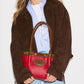 A woman wearing a brown suede jacket, light blue button-down shirt, and light wash jeans holds a red leather handbag with brown leather handles and trim. The bag features a gold-tone embossed logo medallion on the front.