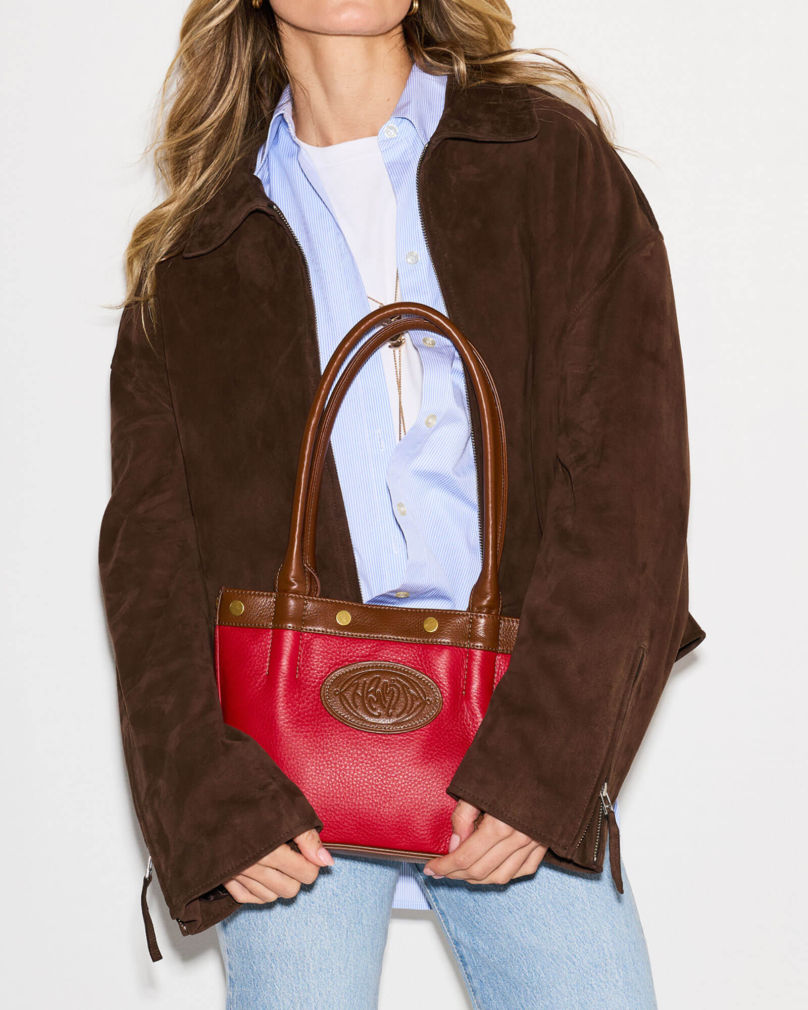 A woman wearing a brown suede jacket, light blue button-down shirt, and light wash jeans holds a red leather handbag with brown leather handles and trim. The bag features a gold-tone embossed logo medallion on the front.