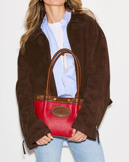 A woman wearing a brown suede jacket, light blue button-down shirt, and light wash jeans holds a red leather handbag with brown leather handles and trim. The bag features a gold-tone embossed logo medallion on the front.