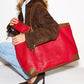 A woman in a brown suede jacket, light blue shirt, and jeans carries a large red leather tote bag with brown leather trim and a matching red pouch.