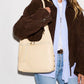 A woman wearing a brown suede jacket, light blue button-down shirt, white tee, and blue jeans carries a large cream-colored leather shoulder bag with gold-tone hardware.