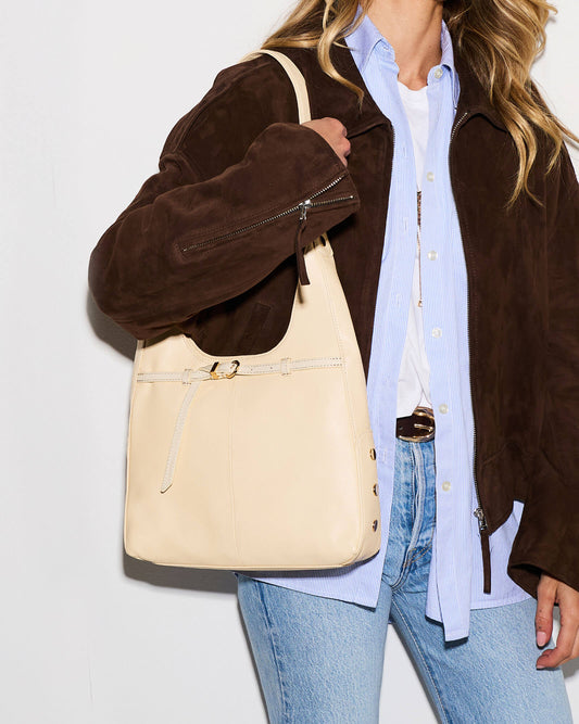 A woman wearing a brown suede jacket, light blue button-down shirt, white tee, and blue jeans carries a large cream-colored leather shoulder bag with gold-tone hardware.
