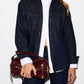 A model wearing a dark indigo denim button-up shirt over a white collared shirt, paired with dark blue jeans and holding a burgundy leather shoulder bag with gold hardware.