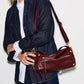 A woman wearing a dark denim shirt and white collared shirt carries a burgundy leather shoulder bag with gold-tone hardware against a plain white background.