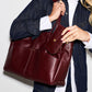 A woman wearing a dark denim jacket and white shirt carries a large burgundy leather tote bag with a matching zippered pouch, shown against a plain light background.