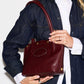 A person wearing a dark denim shirt over a white collared shirt holds a dark burgundy leather shoulder bag with gold-tone hardware against a plain white background.