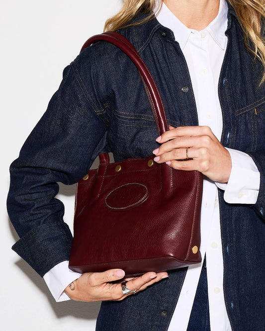 A person wearing a dark denim shirt over a white collared shirt holds a dark burgundy leather shoulder bag with gold-tone hardware against a plain white background.