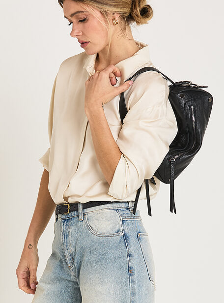 A woman wearing a cream-colored button-up shirt and light blue jeans, carrying a small black leather bag with black hardware over her shoulder against a plain white studio background.