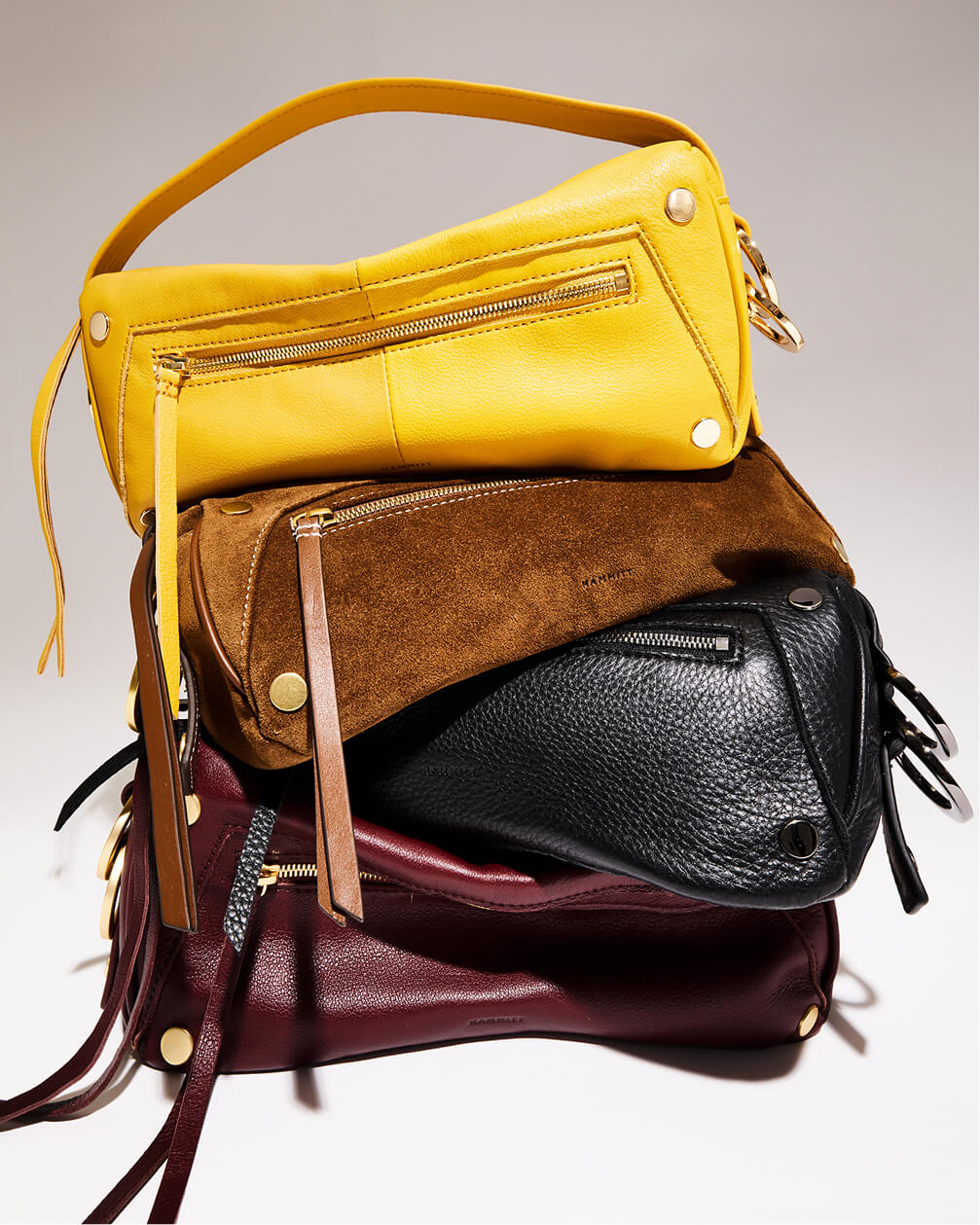 A stack of four designer shoulder bags in yellow, brown, black, and burgundy, featuring textured leather and suede finishes with gold-tone hardware and black hardware.
